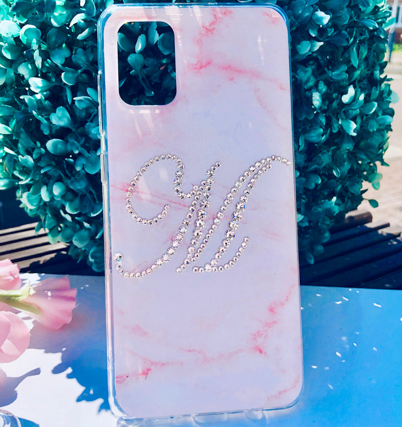 Phone case with pink and white marble design and rhinestone letter 'M' on a blue surface with greenery.